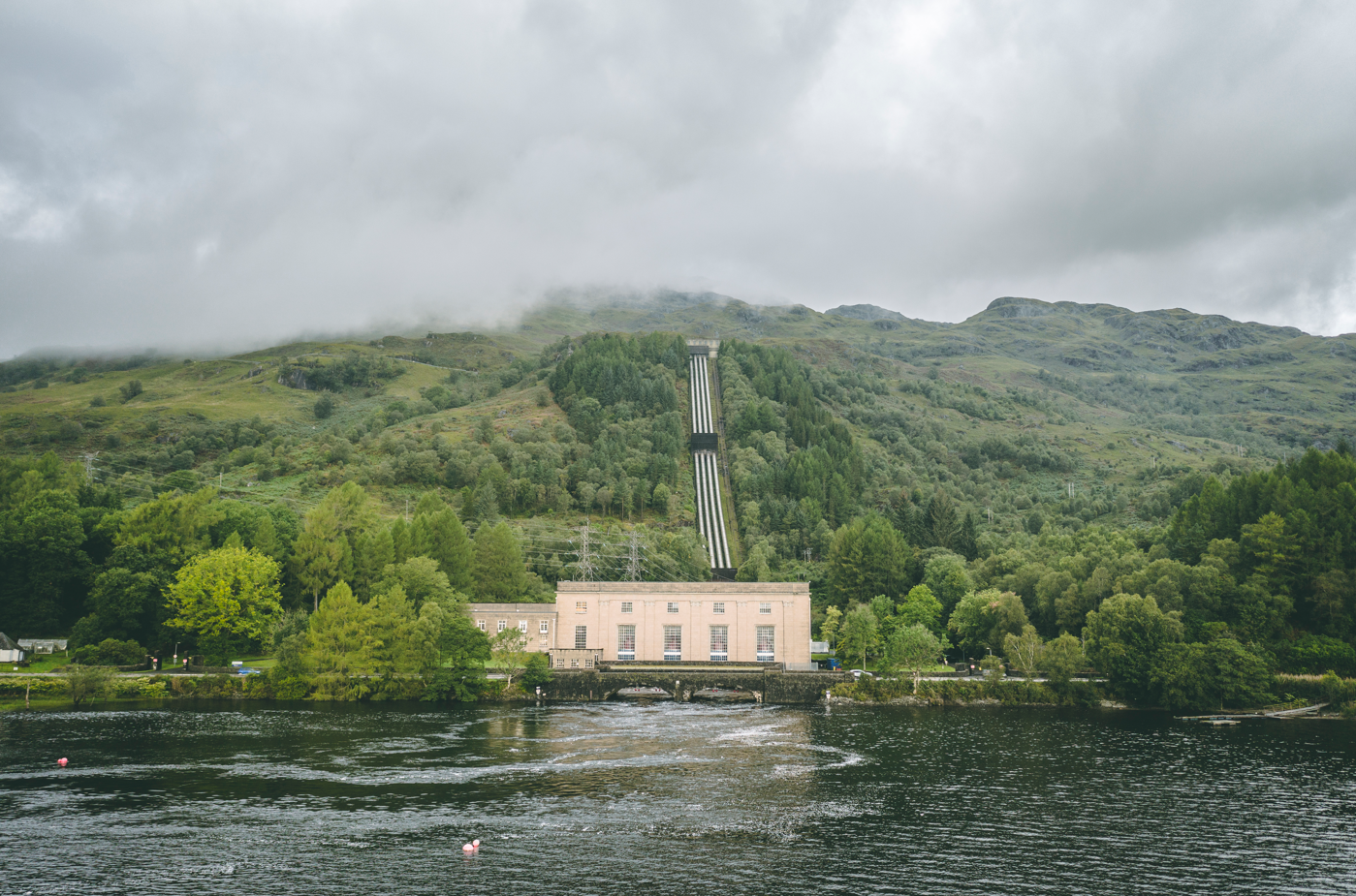 The image depicts a hydroelectric power station located by a serene lake, surrounded by lush green hills under a cloudy sky. The power station features a distinct architectural style with a conveyor system leading up the hillside.