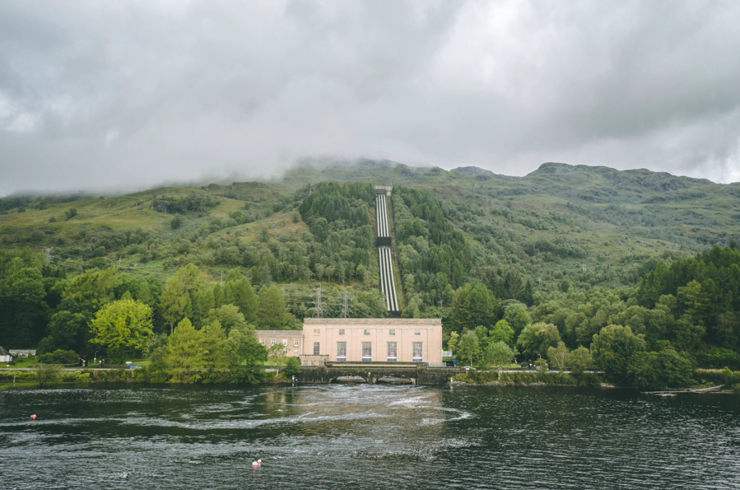 The image depicts a hydroelectric power station located by a serene lake, surrounded by lush green hills under a cloudy sky. The power station features a distinct architectural style with a conveyor system leading up the hillside.