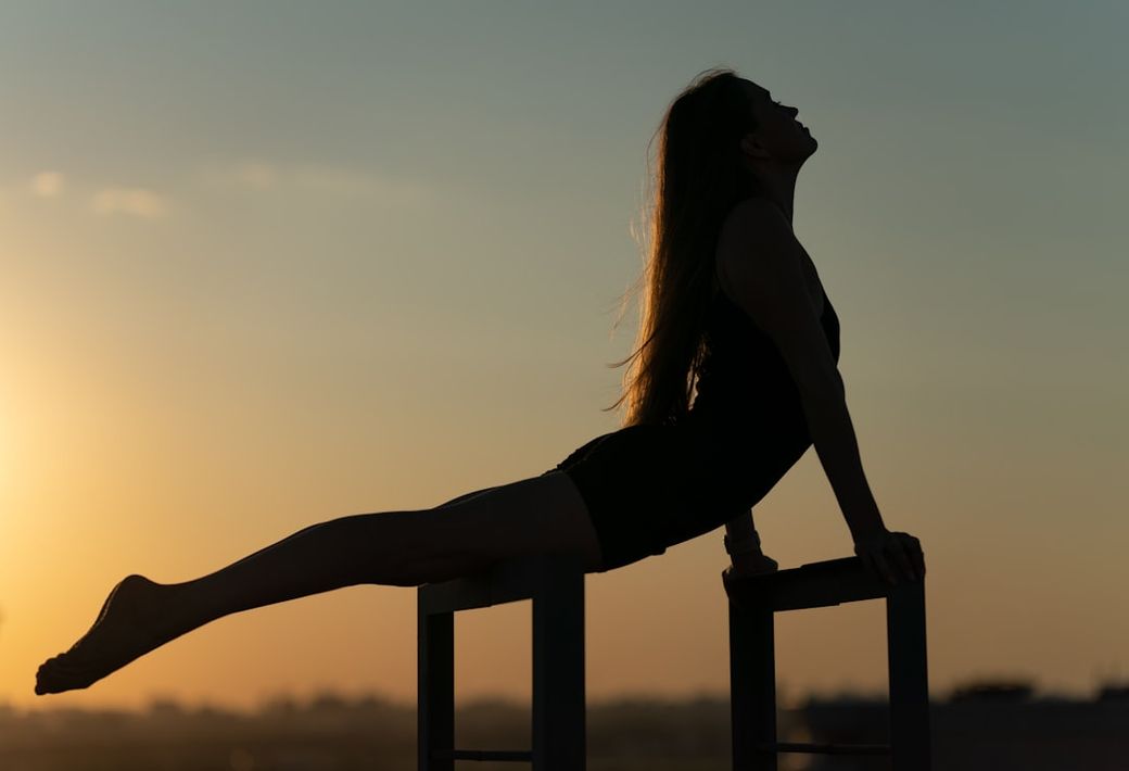 A silhouetted figure performs an acrobatic pose on a chair against a vibrant sunset. The scene captures the beauty of flexibility and balance, highlighting the athlete's strength and grace. — Credit: Photo by Vladyslav Melnyk on Unsplash
