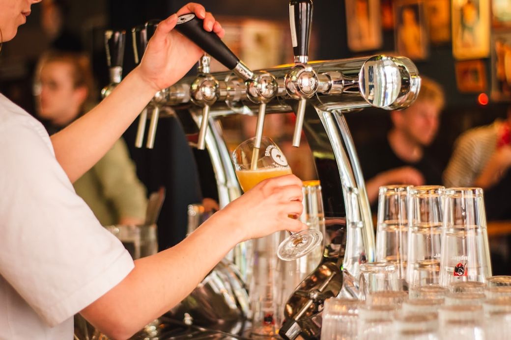 A bartender is serving a draft beer from a shiny tap at a bar. Patrons can be seen enjoying their drinks in the background, creating a lively atmosphere. — Credit: Photo by Louis Hansel on Unsplash