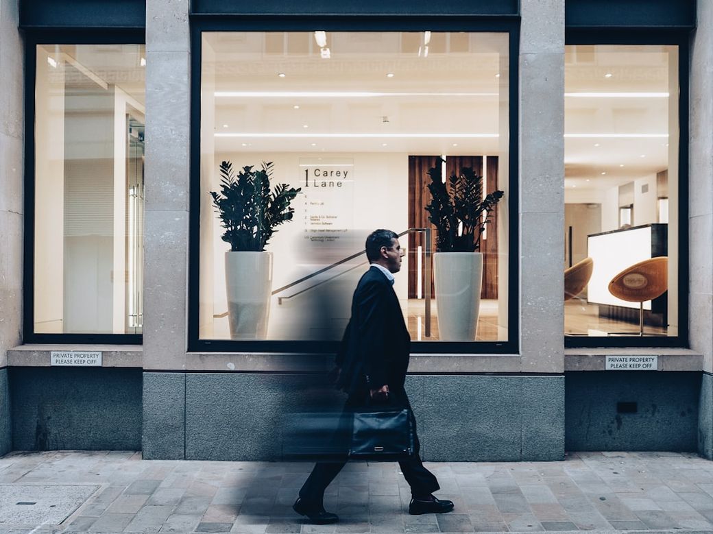 A businessman in a black suit walks past a modern office building with large glass windows. The interior features decorative plants and minimalist furnishings. — Credit: Photo by Nigel Tadyanehondo on Unsplash