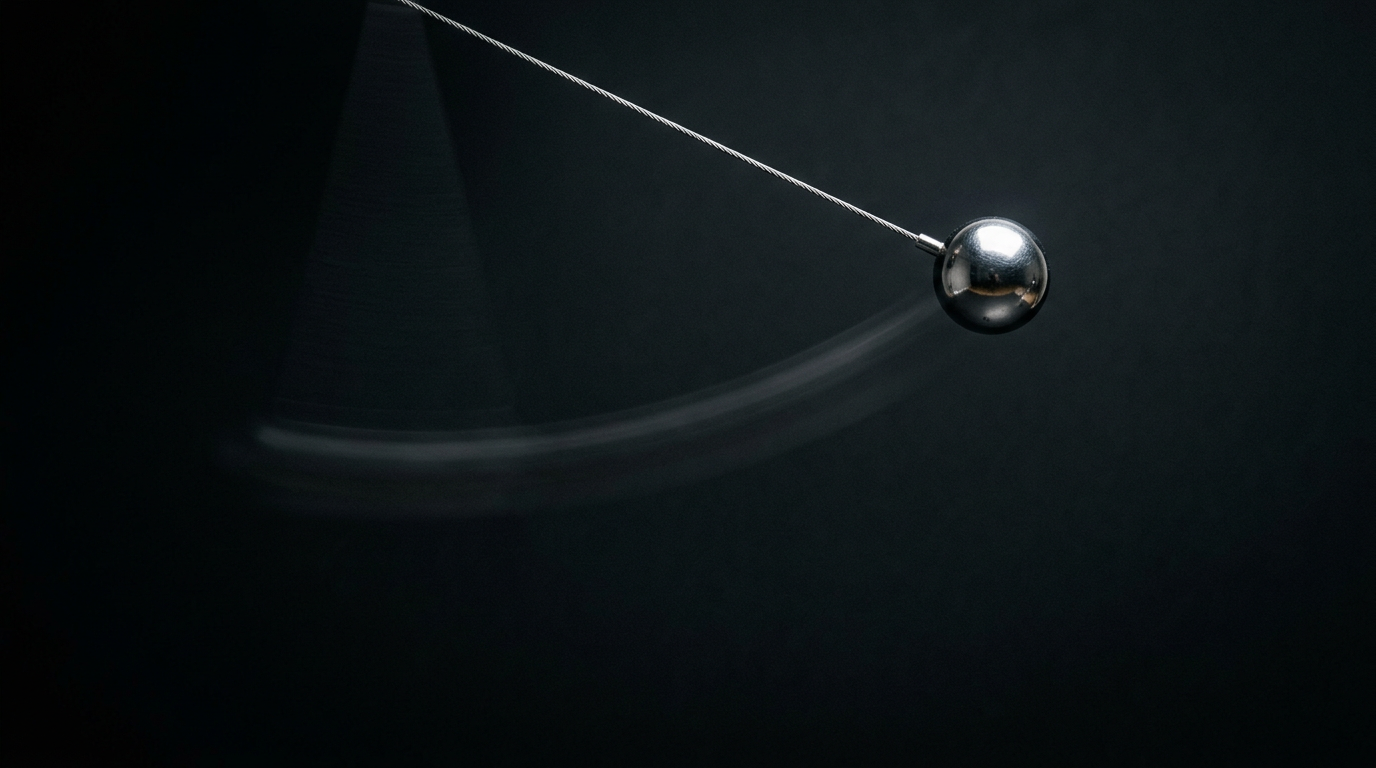 The image captures a polished steel pendulum suspended at the apex of its swing, with a fine wire holding it in place. The dark background enhances the sharpness of the sphere, which is illuminated by a single spotlight, while a faint motion blur indicates its recent movement, creating a striking contrast between stillness and impending motion.