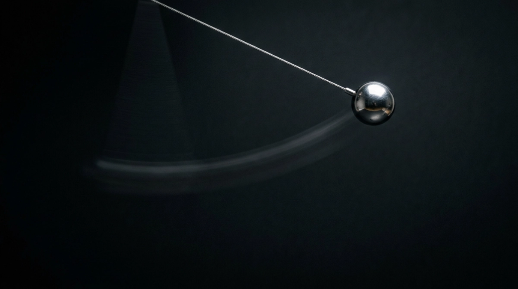 The image captures a polished steel pendulum suspended at the apex of its swing, with a fine wire holding it in place. The dark background enhances the sharpness of the sphere, which is illuminated by a single spotlight, while a faint motion blur indicates its recent movement, creating a striking contrast between stillness and impending motion.