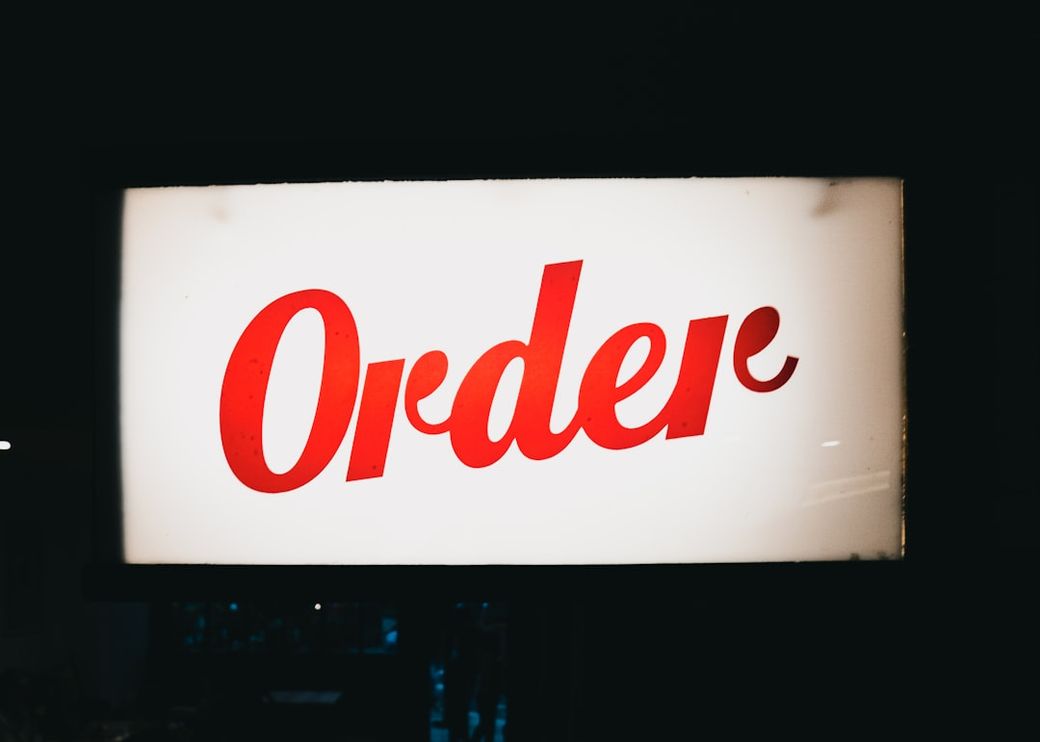 A brightly illuminated sign displaying the word 'Order' in bold red lettering. The sign appears to be part of an indoor setting, possibly in a restaurant or bar, hinting at a place where orders are taken. — Credit: Photo by Samuel Regan-Asante on Unsplash