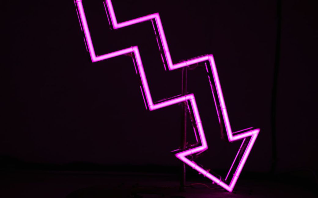 The image features a neon pink zigzag arrow pointing downward, symbolizing a decline or drop. The arrow is illuminated against a dark background, creating a striking visual contrast. — Credit: Photo by Ussama Azam on Unsplash
