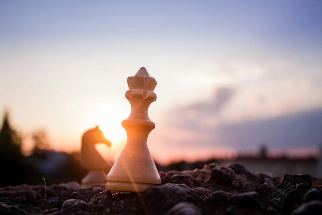 The image features a close-up view of a wooden chess piece, specifically a king, set against a blurred background of another chess piece and a sunset. The warm colors of the sunset provide a dramatic backdrop to the game of chess, highlighting strategy and competition. — Credit: Photo by ᴊᴀᴄʜʏᴍ ᴍɪᴄʜᴀʟ / Unsplash