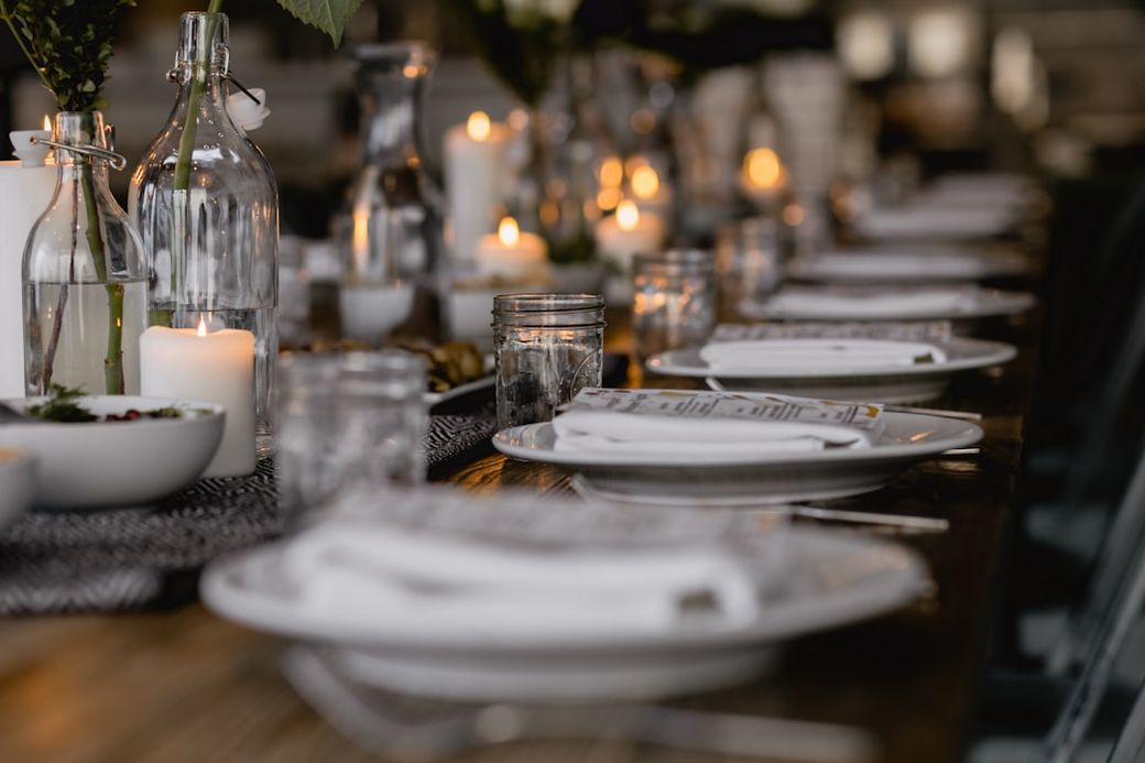 The image depicts a beautifully set dining table with multiple glass containers and candles creating an intimate ambiance. The table is arranged with plates, napkins, and decorative elements, suggesting a special occasion or a formal gathering. — Credit: Photo by Nadia Valko on Unsplash