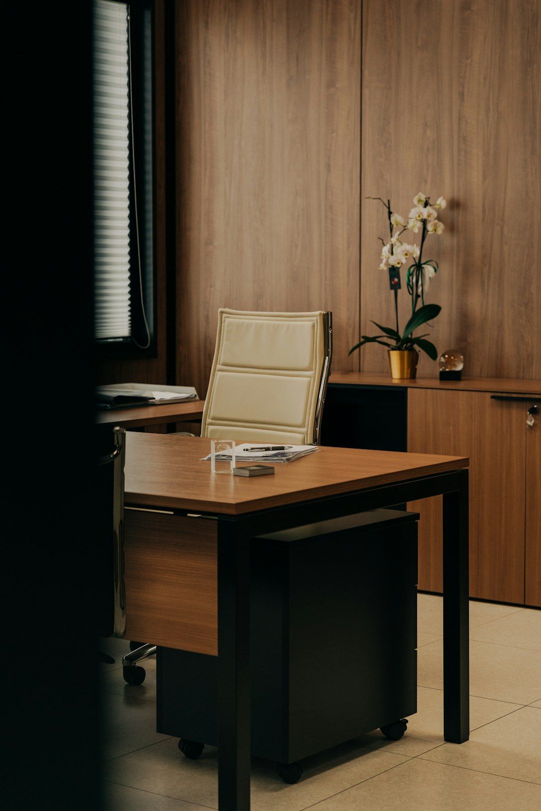 A modern office space featuring a wooden desk and a stylish office chair. The room is adorned with a potted orchid and has a warm, inviting atmosphere.
