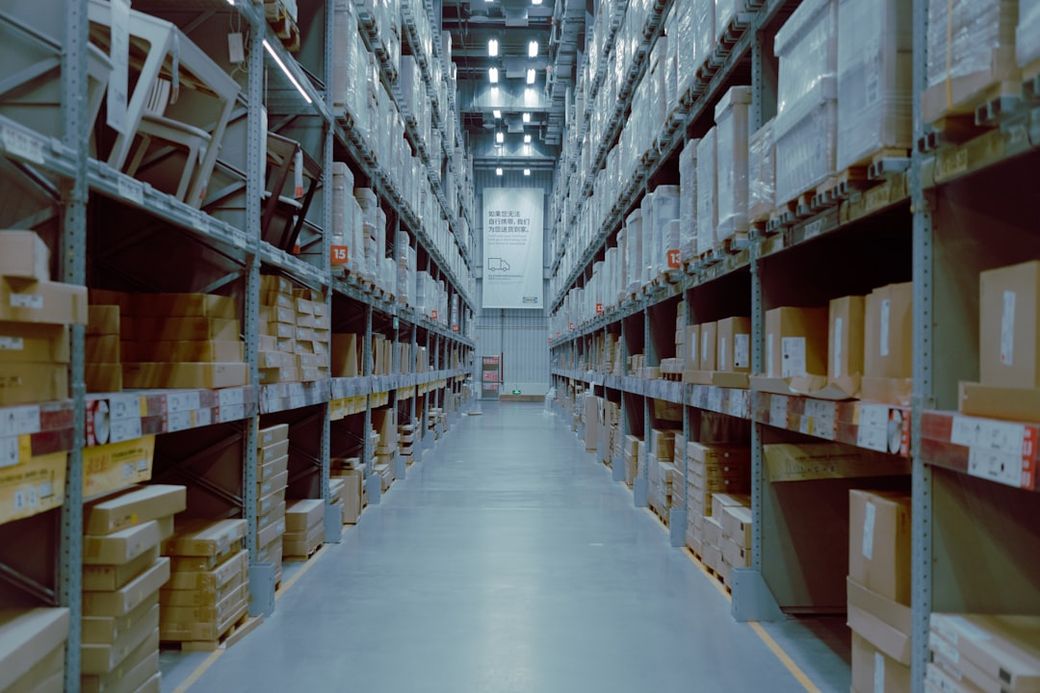 The image depicts a wide aisle within a large warehouse, lined with shelves filled with neatly stacked cardboard boxes. The space is well-lit, highlighting the organized layout of the storage area. — Credit: Photo by Lance Chang on Unsplash