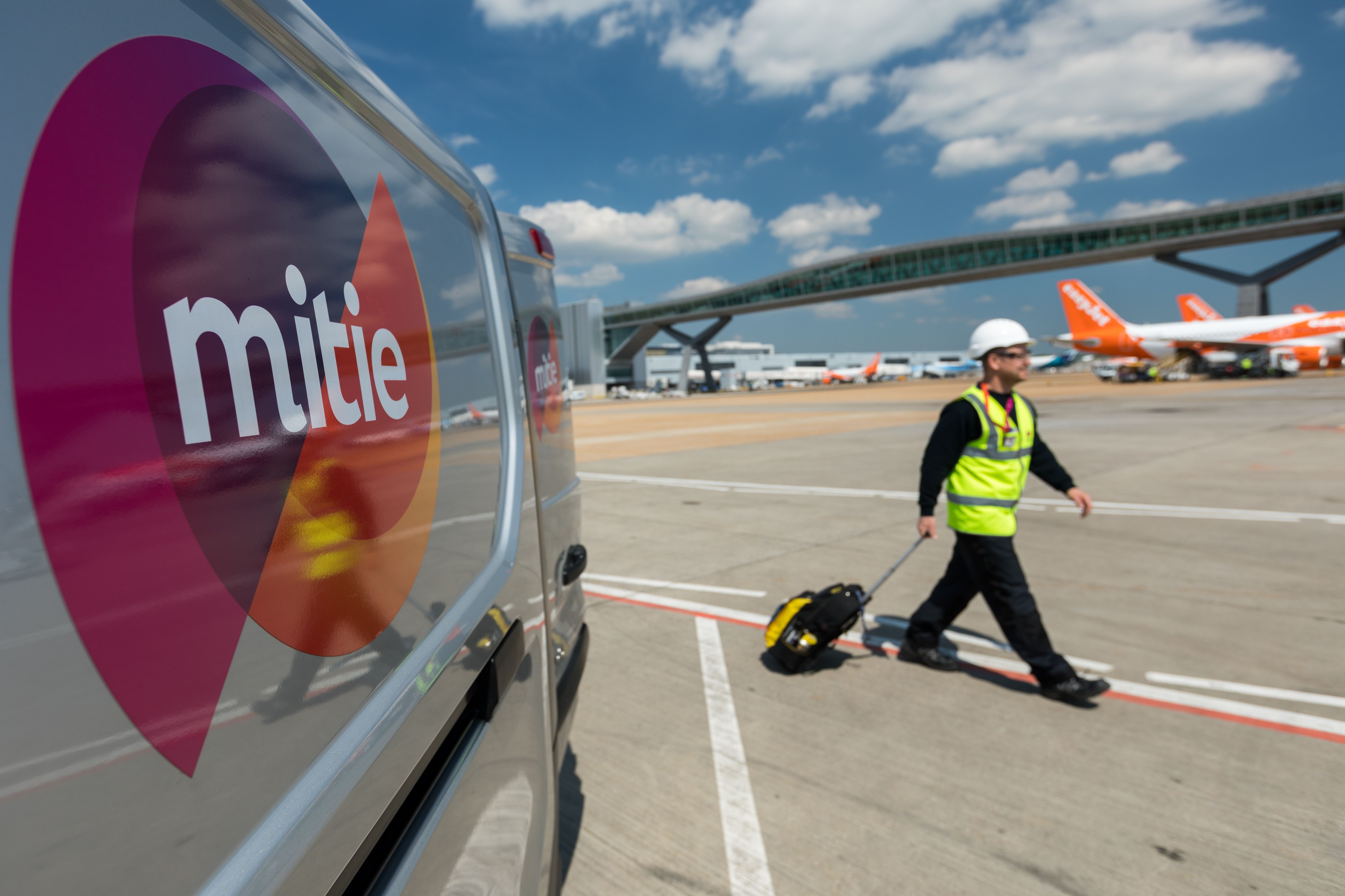 A worker in a hard hat and high-visibility vest walks alongside an airport runway, with a van bearing the Mitie logo in the foreground. In the background, several aircraft are visible under a blue sky with some clouds.