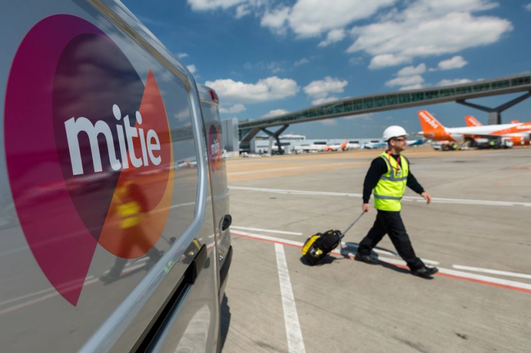 A worker in a hard hat and high-visibility vest walks alongside an airport runway, with a van bearing the Mitie logo in the foreground. In the background, several aircraft are visible under a blue sky with some clouds.