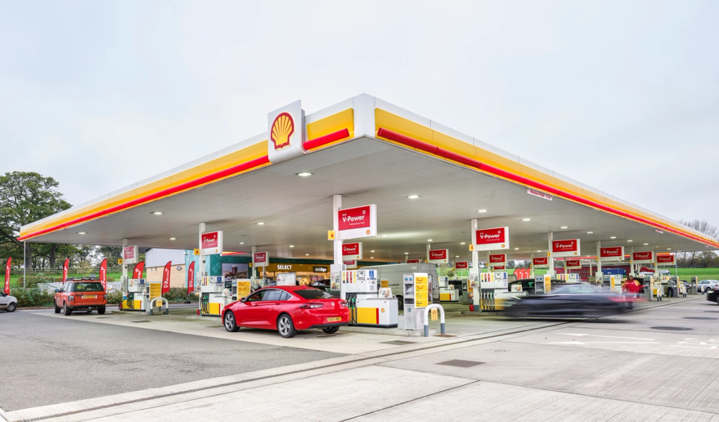 A modern Shell petrol station featuring multiple fuel pumps and a convenience store. Vehicles are seen refueling at the pumps, with the station's distinctive signage prominently displayed.