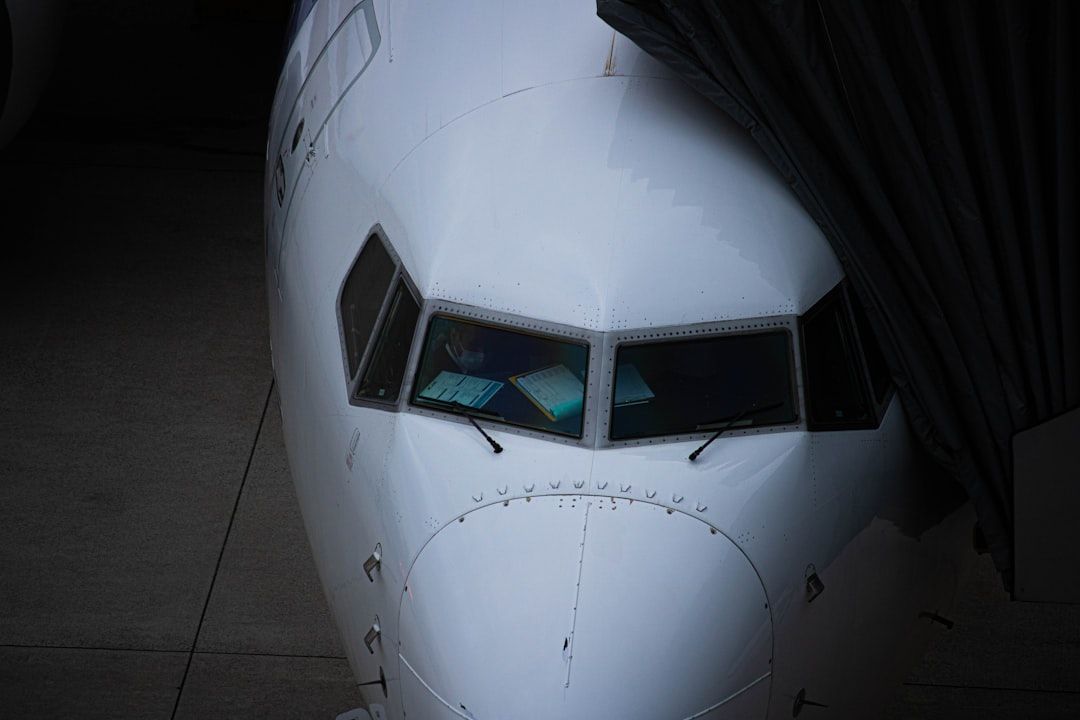The image features the cockpit of a commercial aircraft, showcasing the windshield and the surrounding fuselage. The view is taken from an angle that highlights the folded wing and the surface of the tarmac.