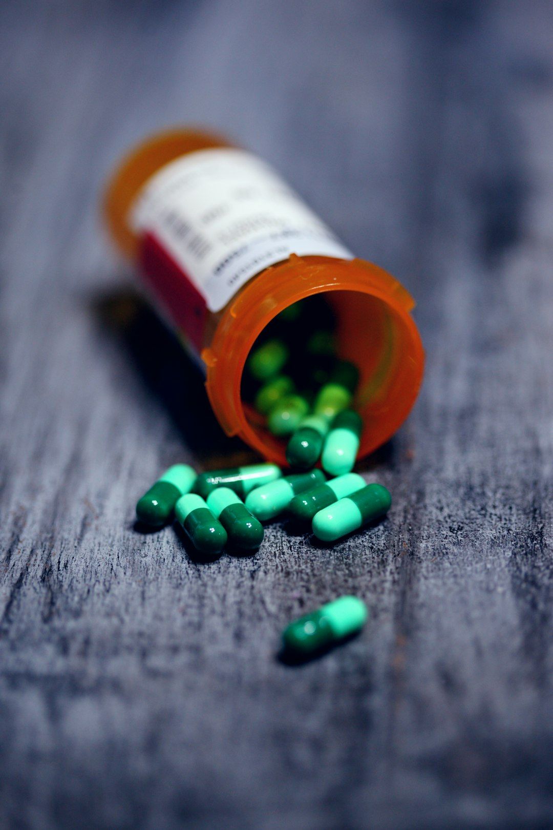 The image shows a prescription bottle that has been tipped over, spilling green and blue capsules onto a dark wooden surface. The focus is on the medication capsules, highlighting their colors and shapes.