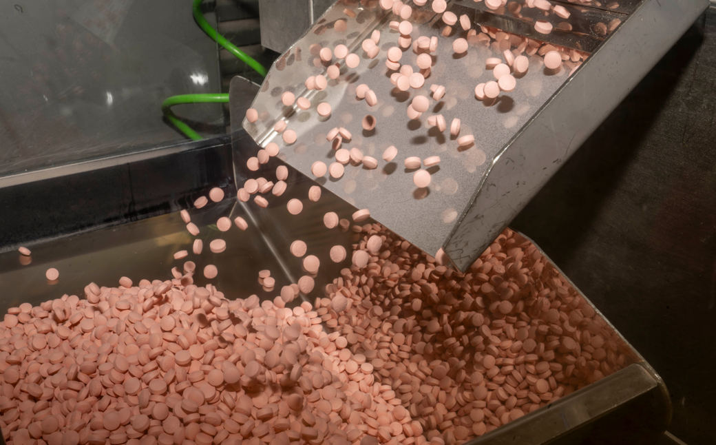 The image depicts a manufacturing process where pink tablets are being poured from a hopper into a large collection tray. The scene illustrates the production of pharmaceutical products.
