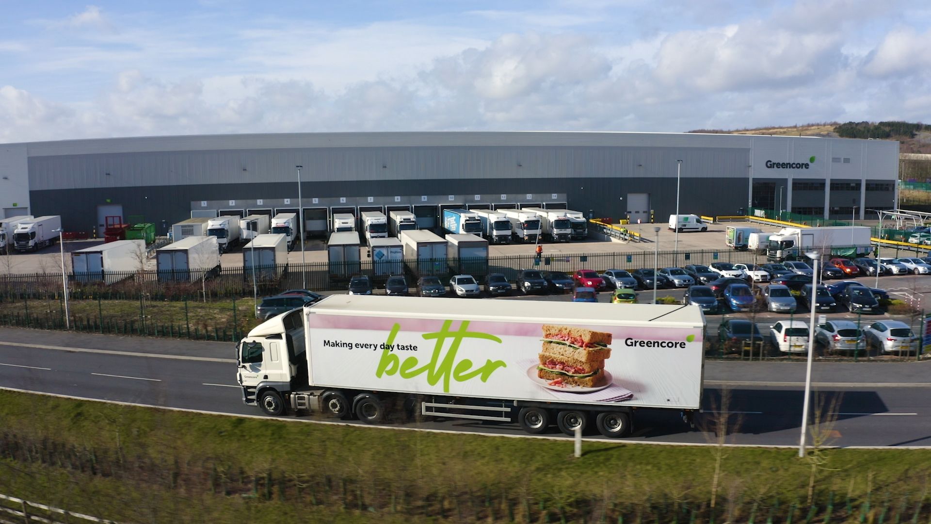 A large truck bearing the logo of Greencore is seen driving past a distribution center. The facility has multiple loading docks and parked vehicles, indicating a busy logistical operation.