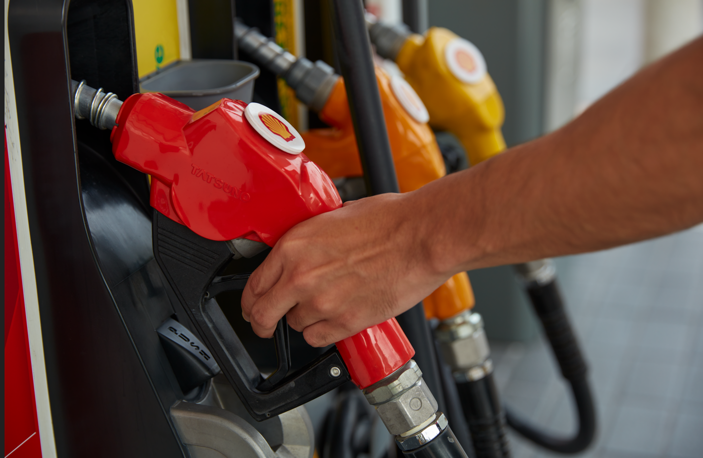 A person holds a red fuel pump nozzle while preparing to refuel a vehicle at a gas station. Various colored fuel pumps are visible in the background, showcasing different fuel options.