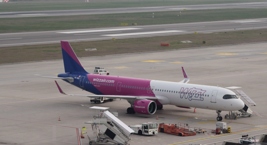 The image shows a Wizz Air aircraft parked on the tarmac at an airport. The plane features a distinctive pink and blue livery, and ground support equipment is visible around it.