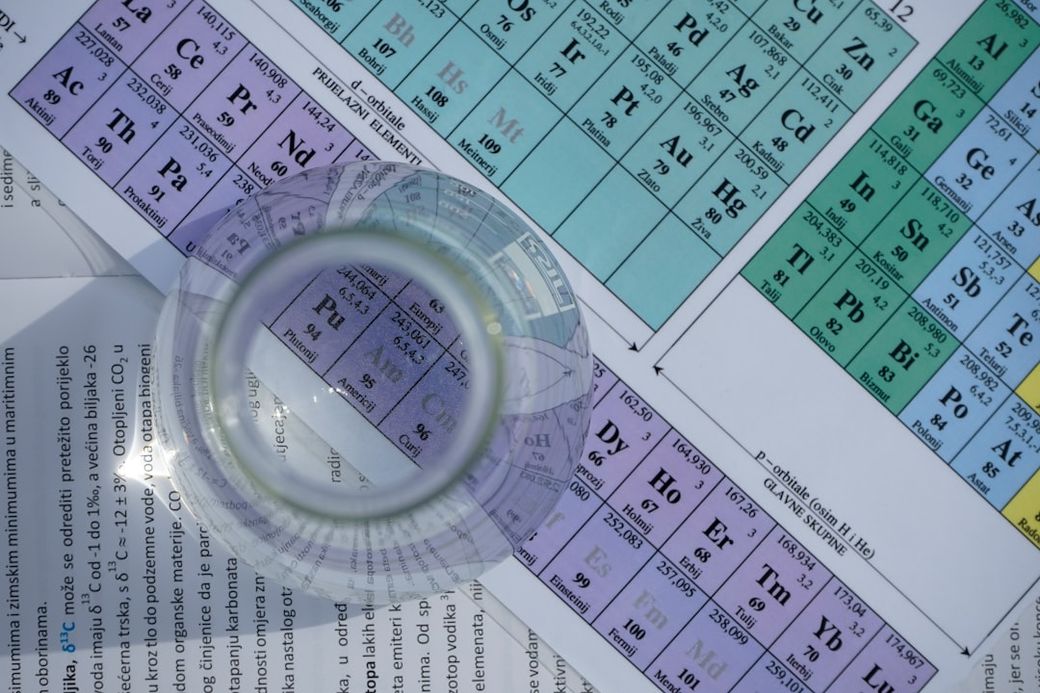 The image shows a close-up view of a clear glass container positioned over a colorful periodic table. The periodic table includes various chemical elements highlighted in distinct shades, suggesting a focus on chemistry education or research. — Credit: Photo by Vedrana Filipović on Unsplash