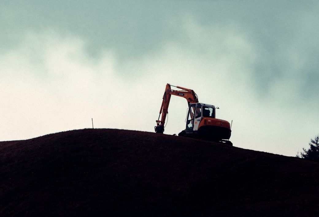 The image features an excavator positioned on a hillside under a cloudy sky. The machinery is silhouetted against the backdrop, emphasizing its size and industrial function. — Credit: Photo by Tobias Kleeb on Unsplash