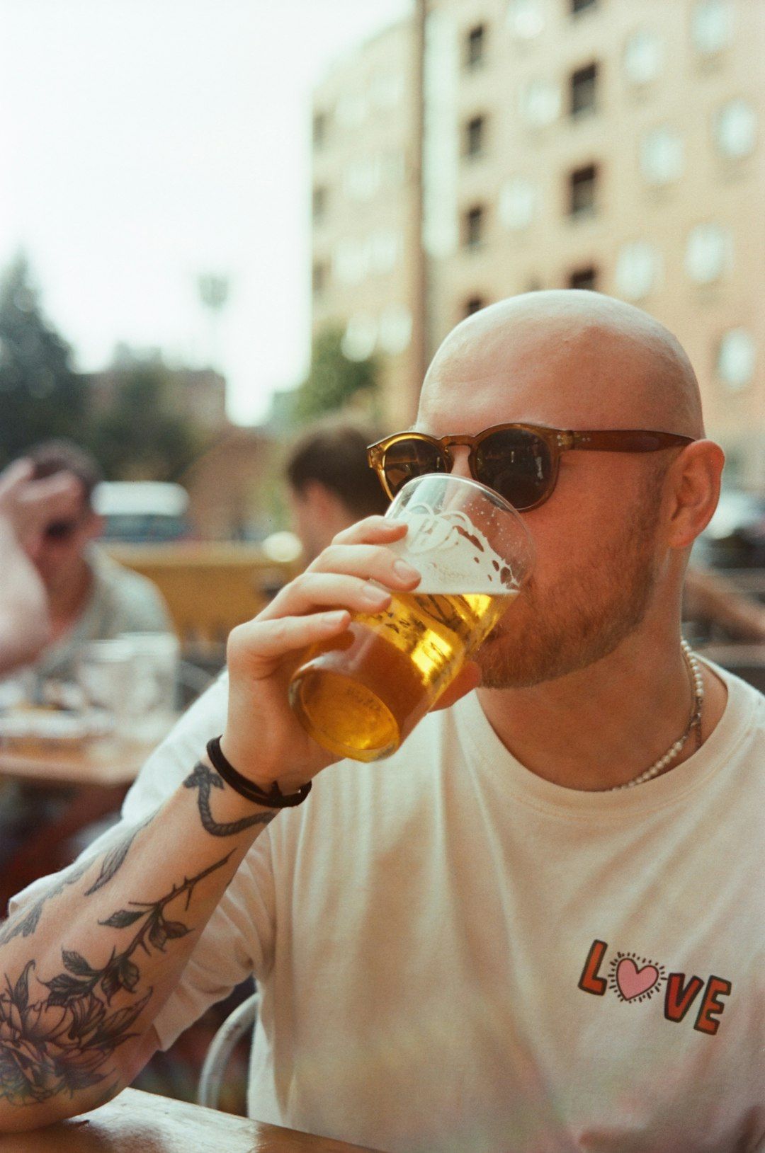 A man with a shaved head wearing sunglasses enjoys a pint of beer in an outdoor setting. The background features partially visible people and buildings, suggesting a casual social atmosphere.
