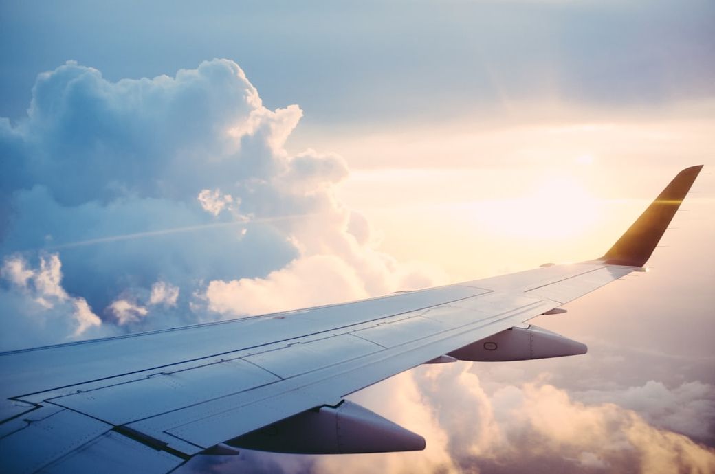 The image depicts the wing of an airplane with a view of fluffy clouds and a colorful sunset in the background. The scene captures the serenity and beauty of flying above the clouds. — Credit: Photo by Ross Parmly on Unsplash