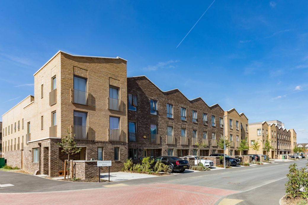 The image depicts a modern residential street featuring newly constructed townhouses with a mix of brick façades. The setting is sunny with clear blue skies, showcasing a well-planned neighborhood.