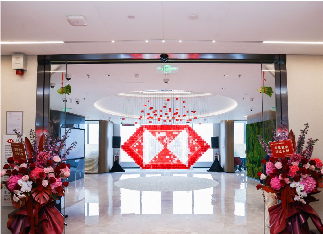 The image depicts a modern entrance area featuring a large decorative installation made of red and white elements. The space is adorned with floral arrangements and has an open, inviting layout with high ceilings.