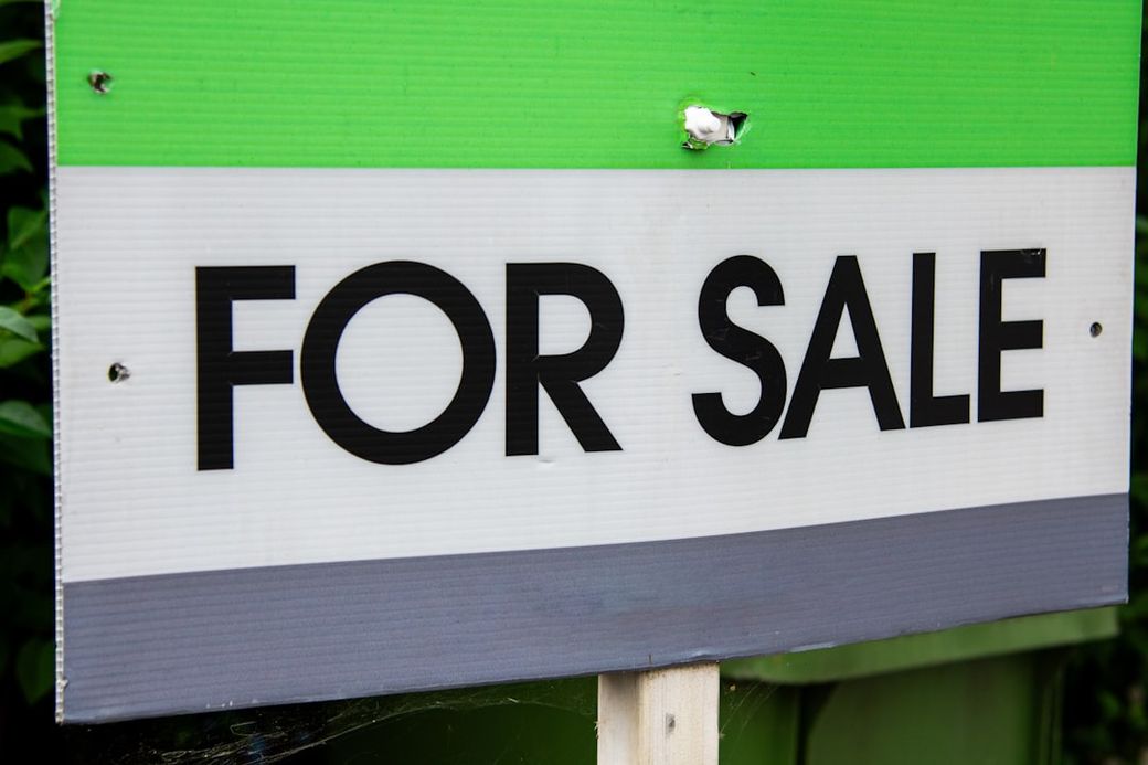 The image features a close-up view of a green and gray 'FOR SALE' sign, indicating a property or item is available for purchase. The sign is prominently displayed with bold lettering against a colored background. — Credit: Photo by Richard Bell on Unsplash