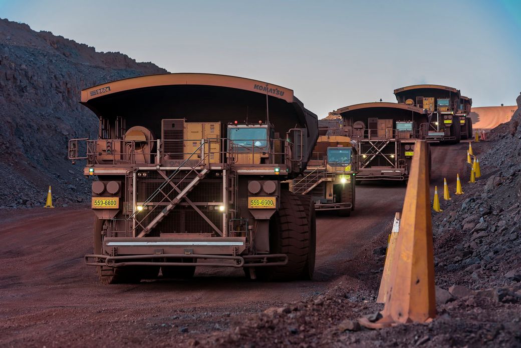 The image showcases large mining trucks operating in a quarry at dusk, surrounded by rugged terrain. The trucks, equipped for heavy-duty tasks, are lined up on a dirt road, highlighting the scale of operations in the mining industry.