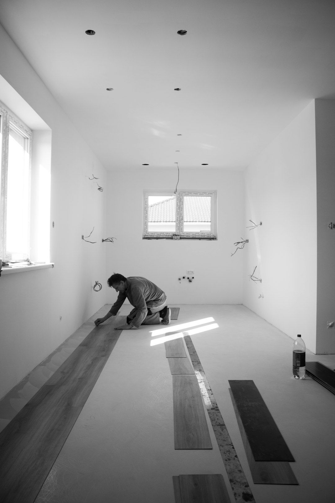 A person is installing wooden flooring in a spacious, empty room with natural light coming through the windows. The walls are bare and the environment appears to be in the middle of renovation.