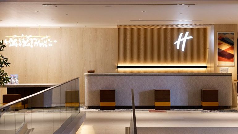 The image features a modern reception area with a sleek design, characterized by a wooden panel accent wall and contemporary lighting. The check-in desk is minimalist, and there are several seating options visible.