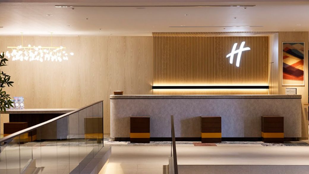 The image features a modern reception area with a sleek design, characterized by a wooden panel accent wall and contemporary lighting. The check-in desk is minimalist, and there are several seating options visible.