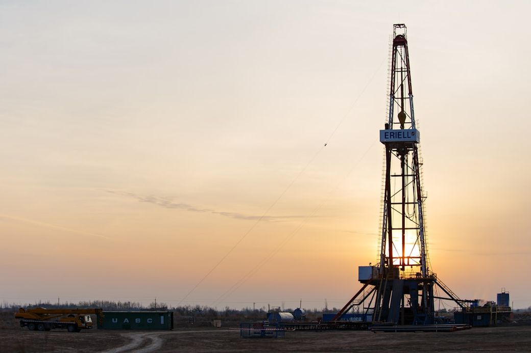 The image captures a drilling rig at sunset, silhouetted against a colorful sky. The rig is located in an industrial area, surrounded by equipment and machinery. — Credit: Photo by WORKSITE Ltd. on Unsplash