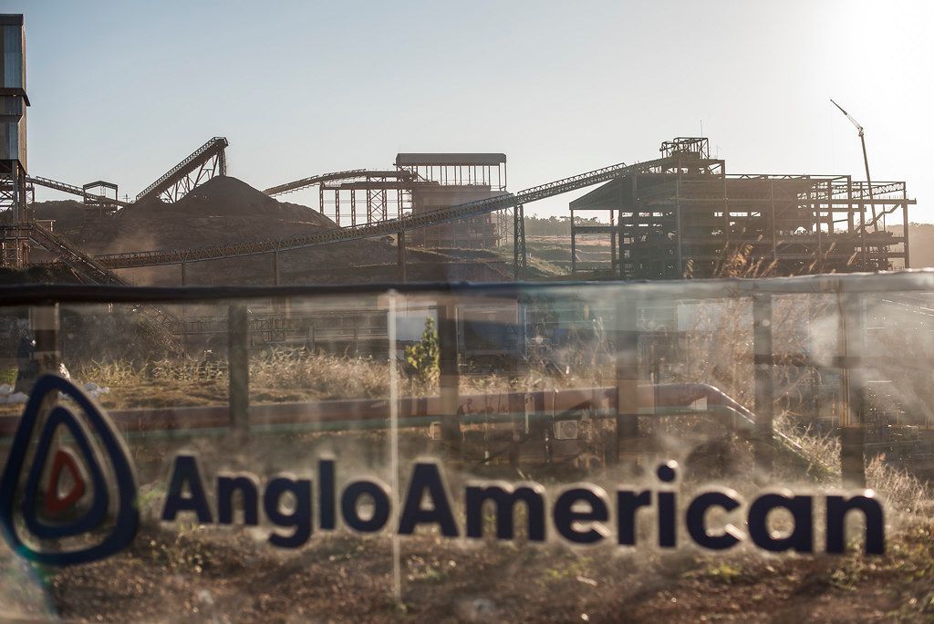 The image showcases an industrial landscape featuring large mining equipment and structures. In the foreground, a clear view of the logo for Anglo American is presented, with piles of material visible in the background under a clear sky.