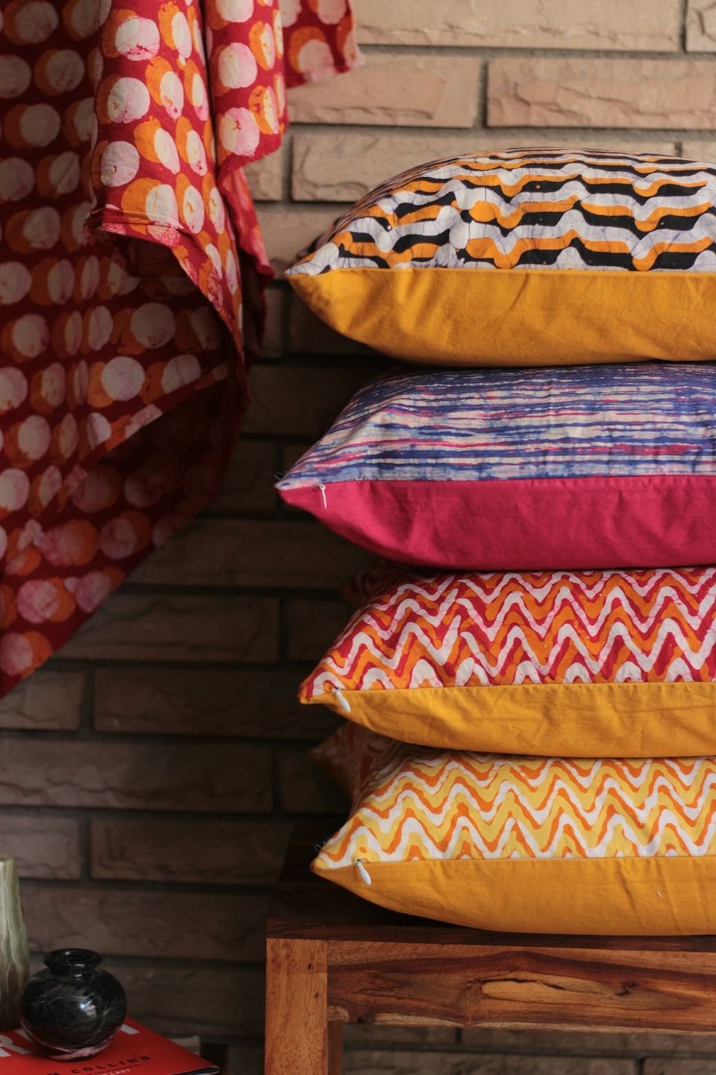 The image features a stack of decorative pillows displayed on a wooden bench. The pillows showcase various vibrant patterns in warm colors, against a textured brick wall, enhancing a cozy atmosphere. — Credit: Photo by Awesome Sauce Creative on Unsplash