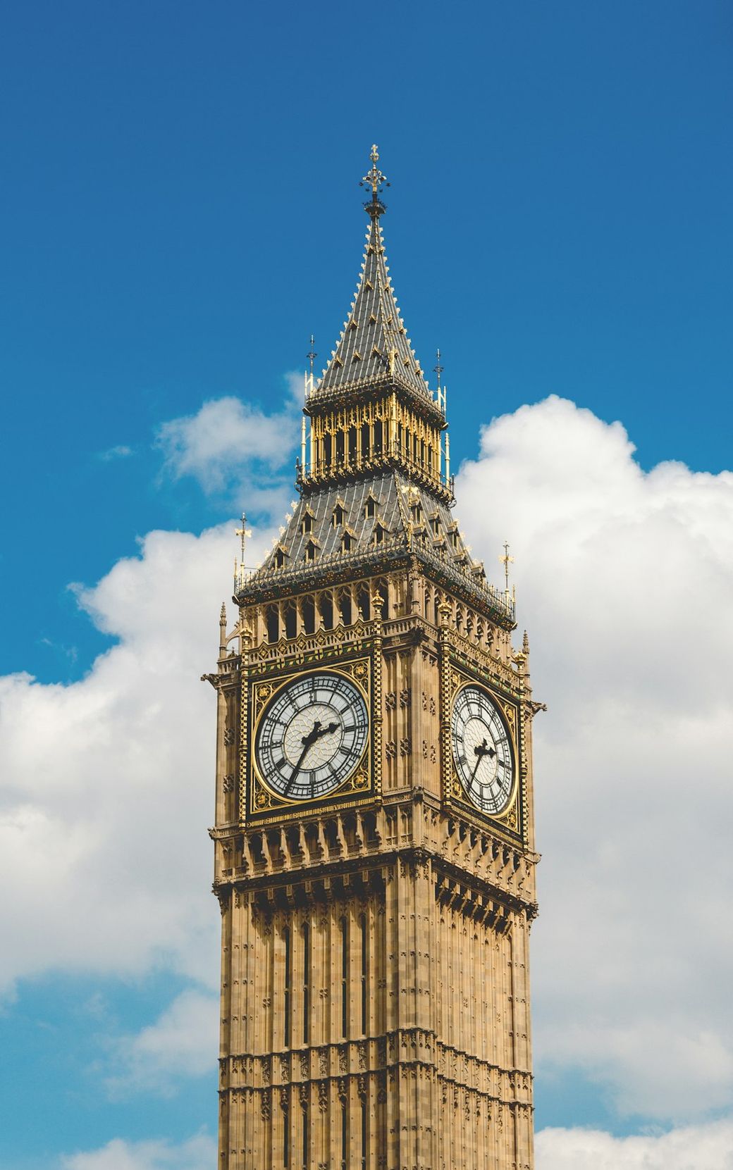 A close-up view of the iconic Big Ben clock tower in London, showcasing its intricate architecture and detailed clock faces. The tower is set against a bright blue sky with a few scattered clouds. — Credit: Photo by enrico bet on Unsplash