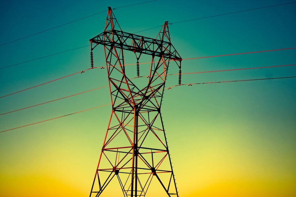 The image shows a high-voltage power transmission tower silhouetted against a colorful sunset. The tower features distinct geometric shapes and is part of the electrical grid infrastructure. — Credit: Photo by Ernest Brillo on Unsplash