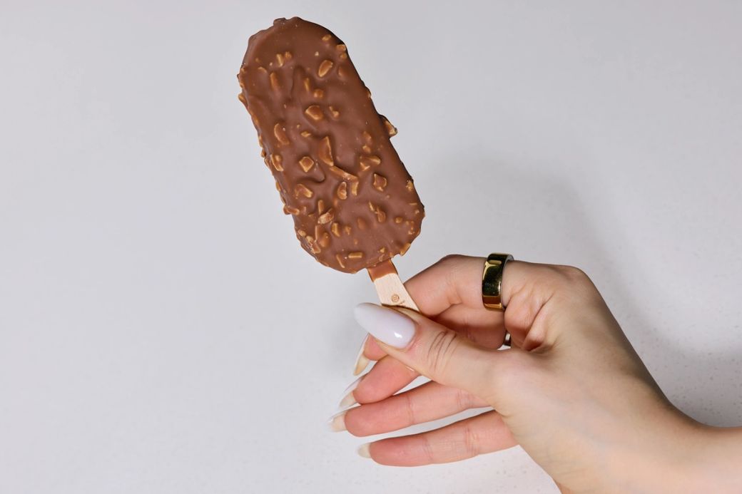 A hand holds a chocolate-coated ice cream bar with nuts on its surface. The background is plain and minimalistic, emphasizing the ice cream treat.