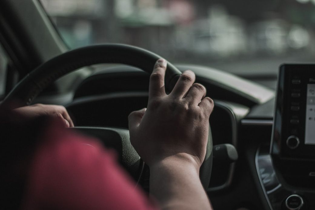 The image shows a close-up view of a person's hand gripping the steering wheel of a car. The interior of the vehicle is partially visible, with a focus on the dashboard. — Credit: Photo by Mae Dulay on Unsplash