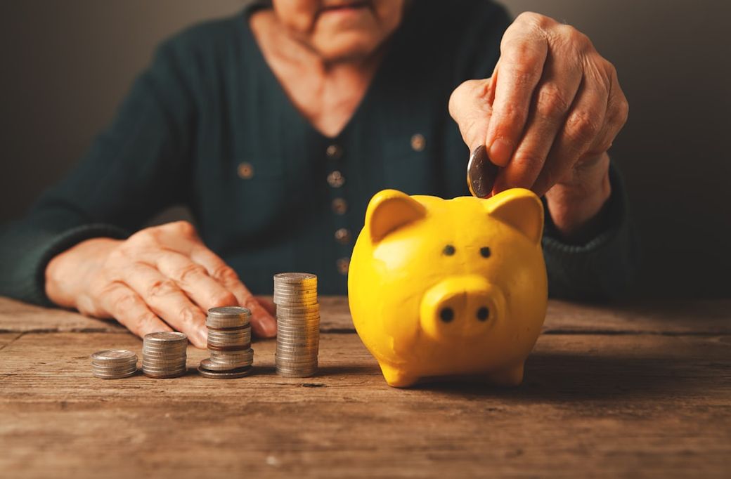 An elderly person is seen placing a coin into a yellow piggy bank, symbolizing savings and financial planning. Stacks of coins are visible in the foreground, highlighting the theme of money management. — Credit: Photo by Sasun Bughdaryan on Unsplash