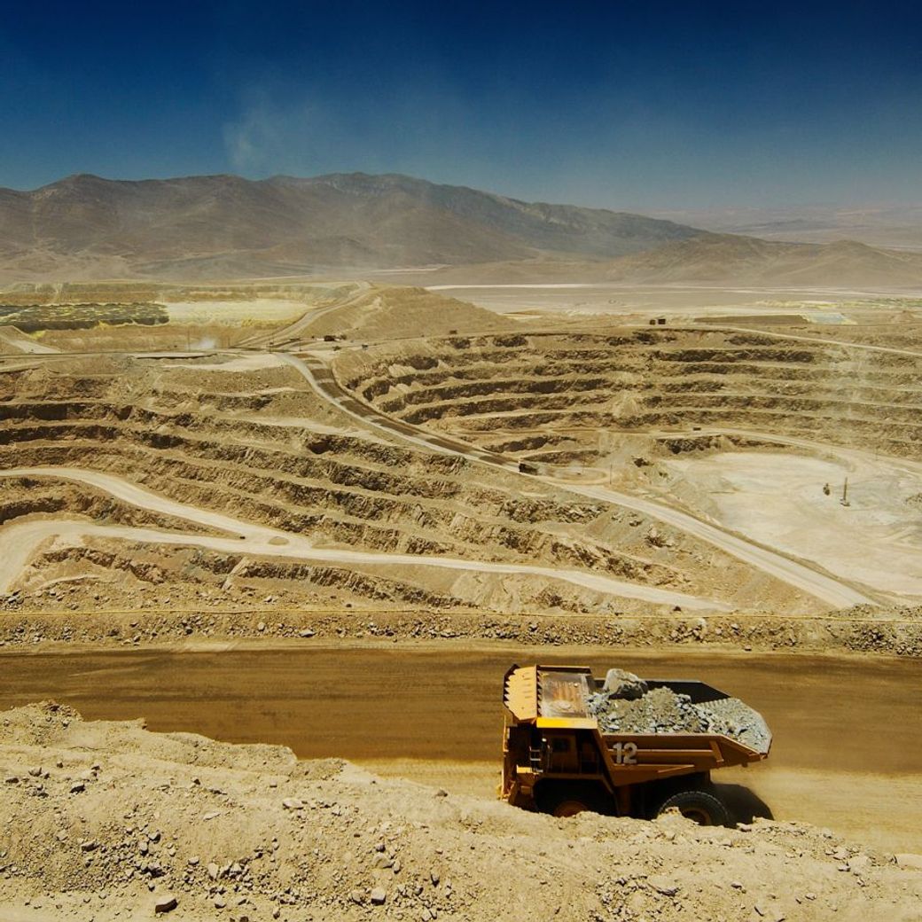 The image depicts a large mining truck on a dirt road within a vast open-pit mine. The surrounding landscape features multiple terraces of excavated earth and a distant mountainous backdrop under a clear blue sky.