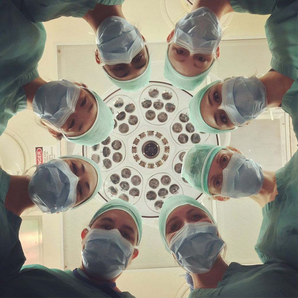 A group of eight medical professionals, dressed in surgical scrubs and masks, are gathered in a circular formation. They are positioned around a surgical light, suggesting an operating room environment. — Credit: Photo by National Cancer Institute on Unsplash