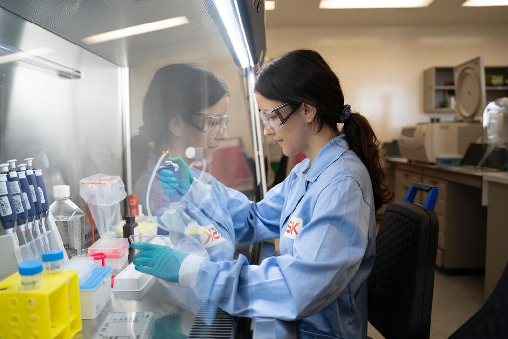 A laboratory technician is performing an analysis in a lab setting, wearing protective gear and using pipettes. The workspace features various laboratory equipment and samples, emphasizing a research environment.