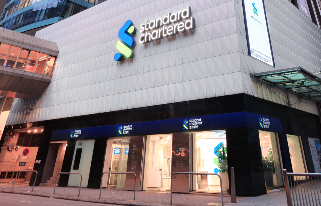 The image shows the exterior of a Standard Chartered bank branch featuring a modern architectural design. The building is well-lit and has a sleek facade, showcasing the bank's logo and signage.