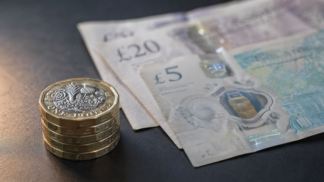 The image features a stack of gold pound coins next to British banknotes, including a £5 and a £20 note. The focus is on the currency, highlighting the details of the coins and the design of the notes.