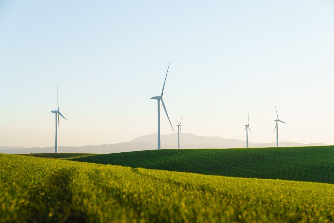 The image depicts a scenic landscape with several wind turbines on rolling green hills under a clear sky. The turbines are set against a backdrop of distant mountains, showcasing a renewable energy installation.