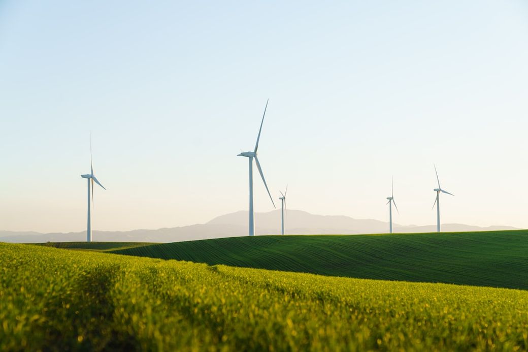 The image depicts a scenic landscape with several wind turbines on rolling green hills under a clear sky. The turbines are set against a backdrop of distant mountains, showcasing a renewable energy installation. — Credit: Photo by Zac Wolff on Unsplash