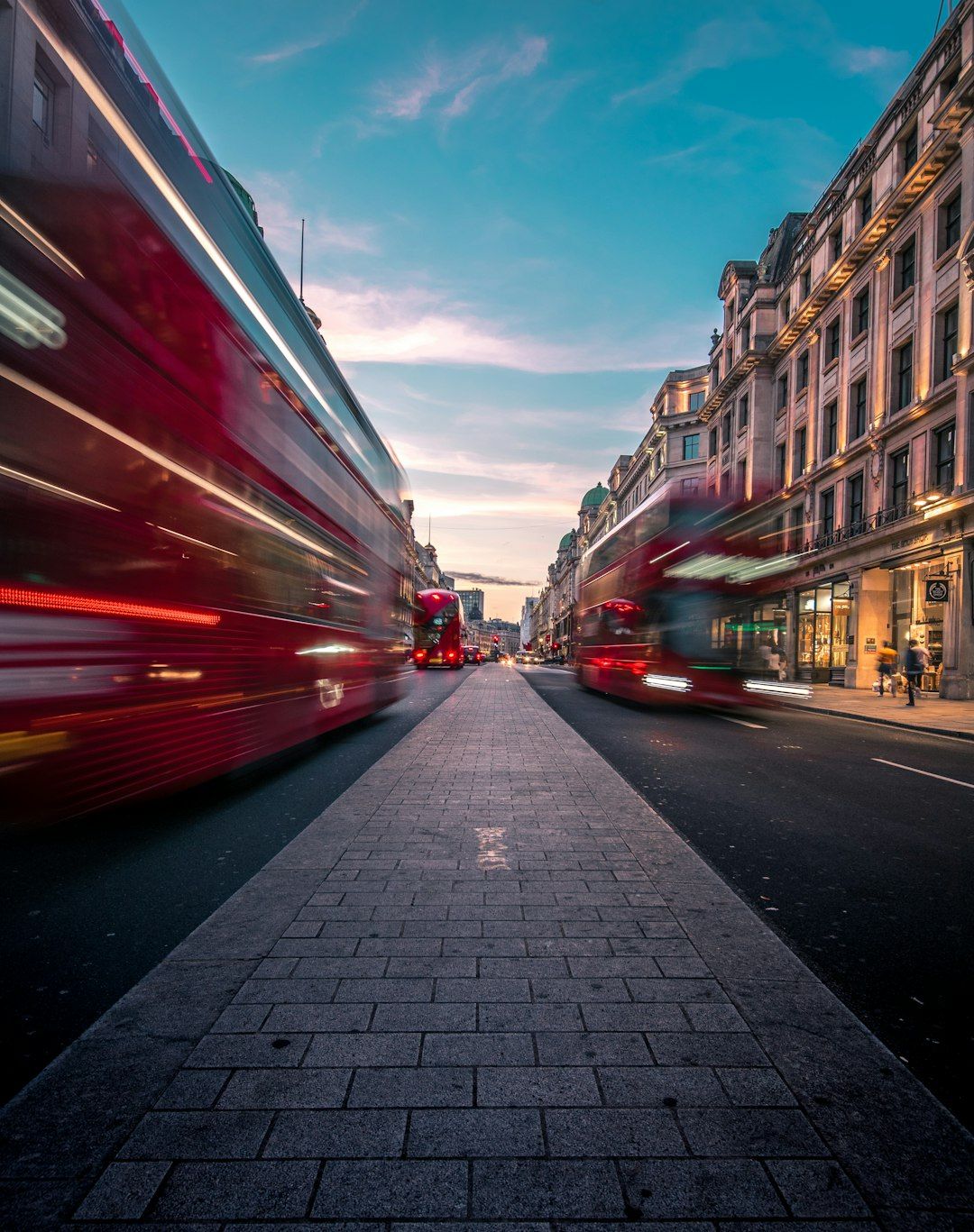 A busy urban street scene featuring iconic red double-decker buses in motion, set against a backdrop of historical buildings and a clear blue sky. The image captures the hustle and bustle of city life with a focus on the street's unique architecture.