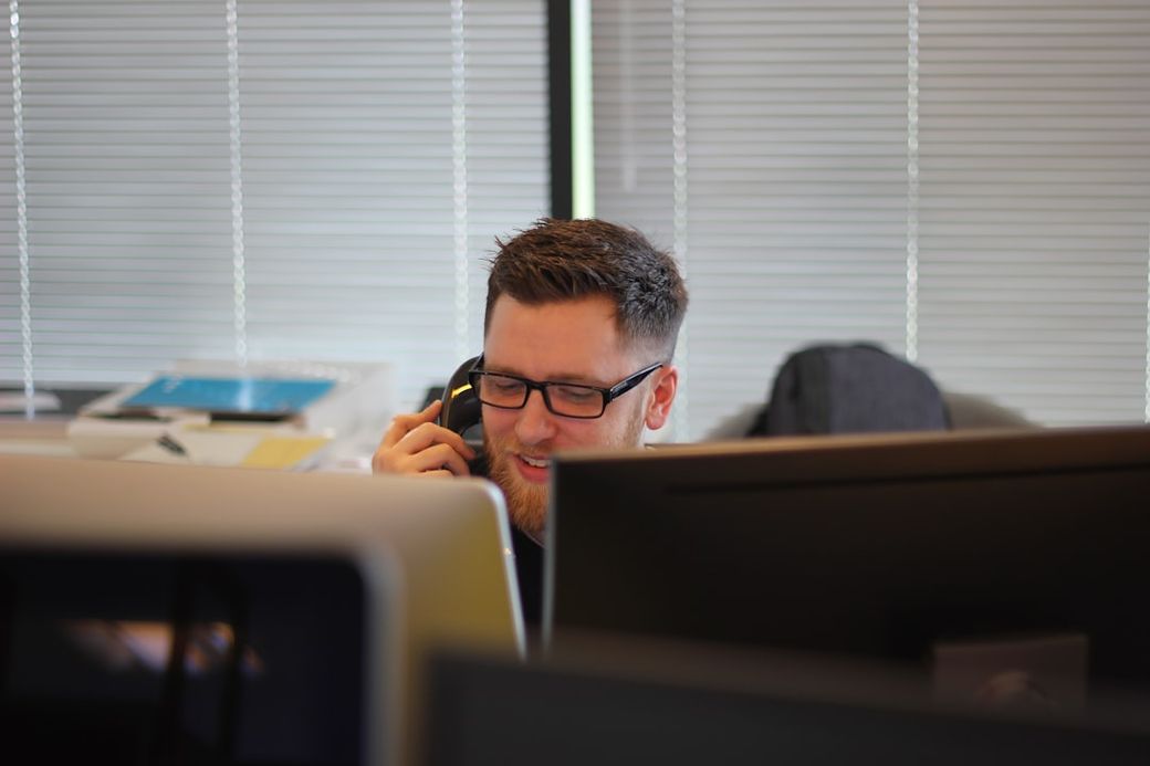 A man is smiling while talking on the phone in an office environment. The background shows multiple computer monitors and office materials, suggesting a busy workplace. — Credit: Photo by Berkeley Communications on Unsplash